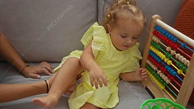 Toddler girl playing with colorful abacus in living room while mother sits nearby