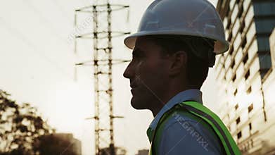 Silhouette of man engineer in safety gear adjusting hardhat standing near electrical transmission lines