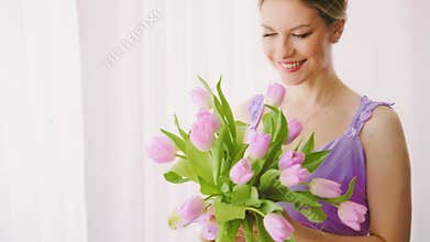 Close-up appy blonde woman holding tulip bouquet purple flowers green leaves white window light. Joyful beauty girl