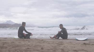 Two surfers relax on the beach, enjoying the sun and waves