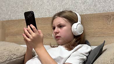 Sad teenager girl with headphones using smartphone lying on bed