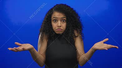 Woman with curly hair and confused expression shrugging against isolated blue background, conveying uncertainty and doubt