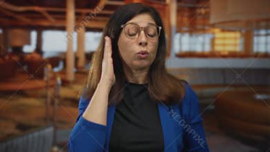 Woman in blue blazer covers ears with fingers in hotel lobby building near seating area; irritation