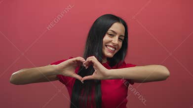 Woman smiling and forming heart shape with hands against red wall, emphasizing happiness, warmth, and love in an isolated setting