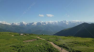 Aerial of person hiking across alpine meadows in mountain range. Hiker walks on trail surrounded by green fields, snowy