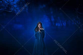 Art Mysterious woman ghost lady. blue dress cape hood. Fantasy queen stands in deep dark mystery forest scared face