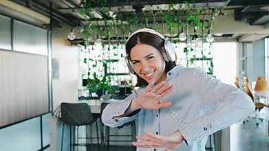 A young woman happily works in a modern office, listening to music with a headset. The office has a comfortable