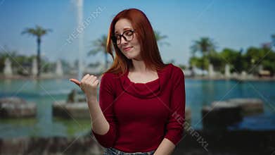 Woman with glasses and red hair in a park, wearing a red sweater, looks amused and points sideways with thumb against a backdrop