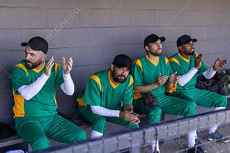 Diverse male teammates sitting on bench in dugout wearing green uniforms, clapping, holding glove