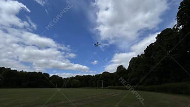 Airliner flying above grassy park with tall trees under bright blue sky