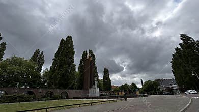 Stormy urban plaza with tall trees, brick monument tower, statues, low wall