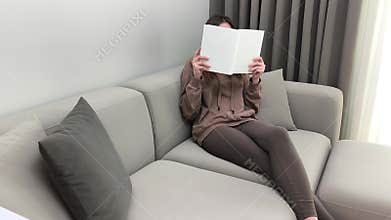 Woman holds book covering face while sitting on sofa. Female