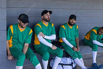 Diverse male teammates sitting on bench in block dugout wearing green jerseys, duffel at feet