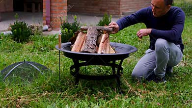 Firewood Arranged by Man in Outdoor Metal Fire Pit Preparing Wood for Lighting Barbecue Fire Near Country House