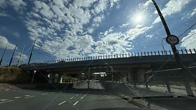 Urban roadway under rail bridge with bright sun, tram wires, traffic signs