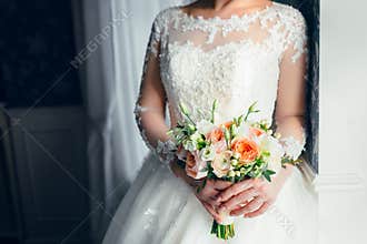 A beautiful bride is standing near the window and holding a wedding bouquet with white roses and peach peonies. Close-up