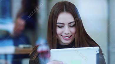 Pretty young woman in a stylish sweater sits in the cafÃ©, drinks coffee, uses the tablet and smiles happily. Cheerful