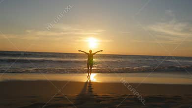 Young woman tourist with backpack walking on beach to the ocean at sunset and raised hands. Girl hiker going on sandy