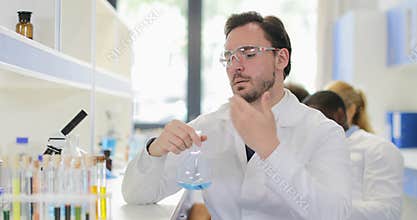 Male Scientist Analyzing Smell Of Liquid In Flask Working In Chemical Laboratory Wear White Coat And Protective Glasses