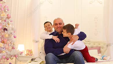 Fun cheerful father and children of twin brothers, on large bed in bright bedroom with Christmas tree.
