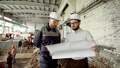 Two men in protective helmets are working together in construction area with workers welding in the background. Foreman
