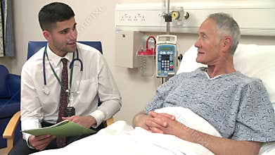 Doctor Sitting By Senior Male Patient's Bed In Hospital