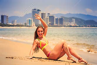 Blonde girl in bikini sits bent knees on wet sand waves hand