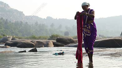 Woman standing at the riverbank and folding long colourful fabric.