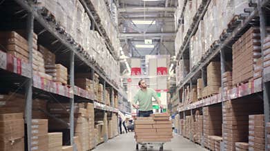 A man is pushing a trolley full of boxes on it between shelves with cardboard boxes in a storage warehouse