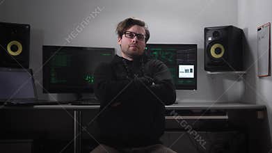 Portrait of a hacker sitting on the chair with computer monitors and loudspeakers behind him. Cyber spy hacked database