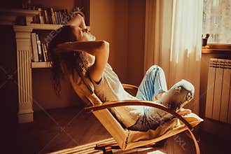 Woman relaxing after work in comfortable modern chair near window in livingroom. Warm natural light. Cozy home. Casual clothing. C