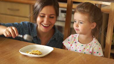 A happy mother is feeding a little daughter. Mom plays a flying rocket. The girl opens her mouth wide. Maternal kiss
