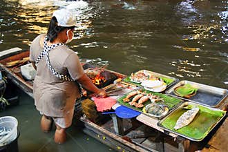 Woman standing in water is cooking seafood for tourists on floating market. Bangkok, Thailand.