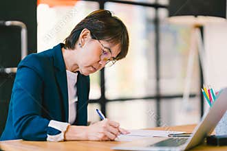 Middle age Asian woman working on paperwork in modern office, with laptop computer. Business owner or entrepreneur concept