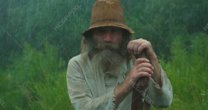 Close-up portrait of the old man with long beard, in old clothes is chewing while leaning on the cane in the rain.