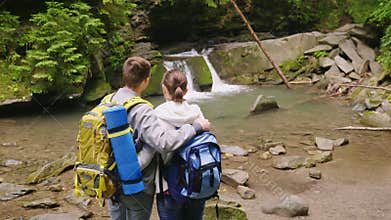 Crane shot: A couple of tourists with backpacks admire the beautiful waterfall and the mountain river. Back view