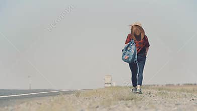 Young stylish girl in cowboy clothes doing hitch-hiking in desert, on the road. She carries backpack and hat. Pass