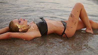 slim model in black leather bikini lying on the shore of the beach in the waves looking in the camera and flirting