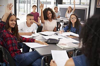 High school kids raise hands, teacher sitting at their desk