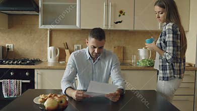 Upset young man reading unpaid bills and hugged by his wife supporting him in the kitchen at home