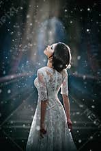 Lovely young lady wearing elegant white dress enjoying the beams of celestial light and snowflakes falling on her face