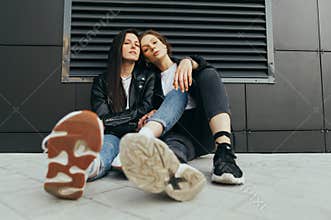 Fashionable photo of two stylish girls in casual clothes wearing leather jackets and sneakers,sitting on the ground against a dark