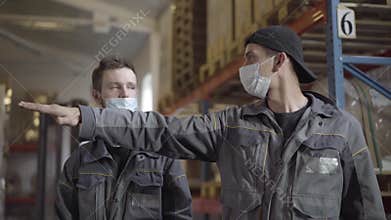Two Caucasian men in face masks discussing work plan in warehouse. Portrait of professional male employees working at