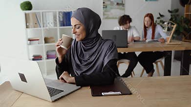 Muslim woman drinking coffee and having a video talk on a laptop in the office
