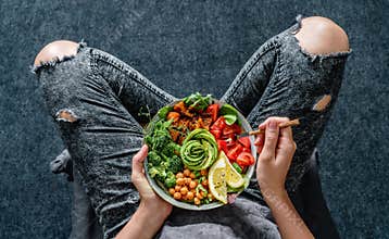 Woman in jeans holding Buddha bowl with salad, baked sweet potatoes, chickpeas, broccoli, greens, avocado, sprouts in hands.