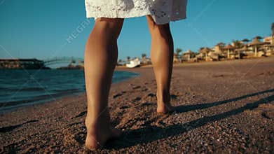Young woman legs walking along sand coastline at summer vacation.
