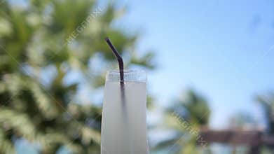 Glass of refreshing summer drink lemonade with plastic straw swaying in sea wind on blurred tropical beach palm trees blue sky