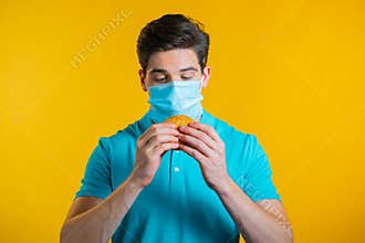 Young man in protective medical mask eating burger on yellow studio background. Guy makes fun, shows how to live during