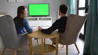 Man and woman are sitting in chairs, kissing and watching TV with a green screen, switching channels with a remote
