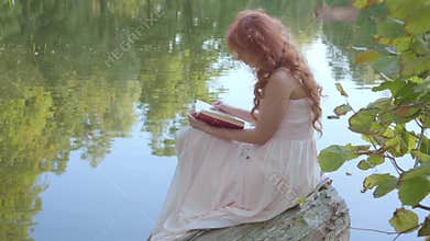 Redhead attractive caucasian girl reading the book in dark red cover on the bank of the lake in the summer forest. Fairy